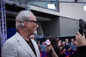 Un homme plus âgé parlant aux journalistes sur un tapis rouge tout en portant un costume prévu d'après David Fincher.