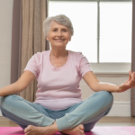 Une femme âgée est assise sur un tapis de yoga rose tout en pratiquant des exercices d'étirement doux.