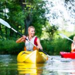 Deux personnes faisant du kayak sur une rivière calme, dont une dans un kayak jaune au premier plan, ressemblant à une automobile en mouvement fluide.