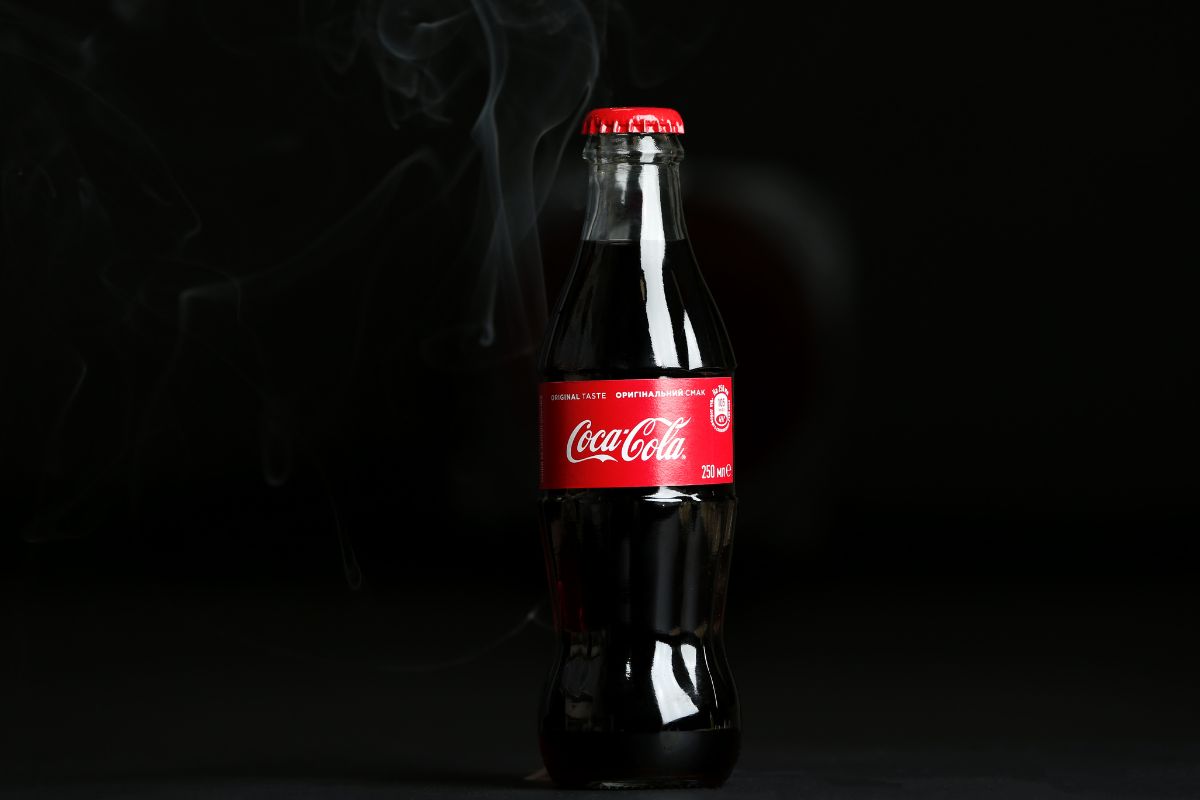 Une bouteille de Coca-Cola en verre avec un bouchon rouge sur fond sombre avec de la fumée alimentaire anti-âge tourbillonnant autour d'elle.