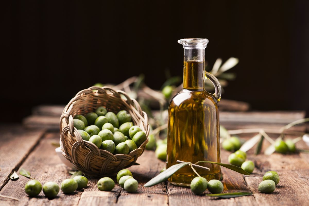 Une bouteille d'huile d'olive, connue pour ses propriétés alimentaires anti-âge, à côté d'un panier d'olives fraîches et de branches d'olivier sur une table en bois.