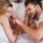 Un couple joyeux interagissant de manière ludique alors qu'il est allongé dans son lit, l'homme touchant le nez de la femme et tous deux se souriant chaleureusement alors qu'ils redécouvrent l'intimité.
