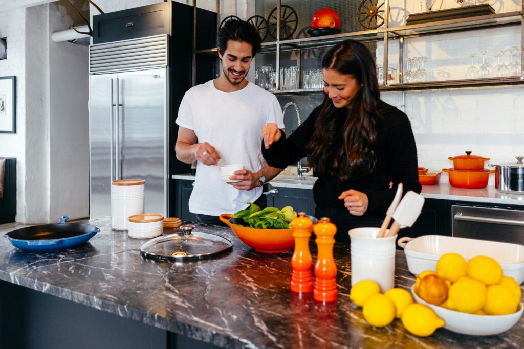 Un jeune homme et une jeune femme souriant et préparant des plats ensemble dans une cuisine moderne avec des ustensiles de cuisine colorés et des ingrédients frais sur le comptoir, utilisant leur auto Brouillon pour les recettes.