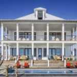 Maison blanche à deux étages dotée de nombreuses fenêtres et colonnes, ainsi que de plusieurs chaises et tables de patio aux deux niveaux. Au premier plan se trouve une piscine avec des chaises longues rouges, ressemblant à une scène d'un auto Brouillon. Ciel bleu clair au-dessus.