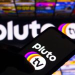 Silhouette d'une main tenant un smartphone affichant le logo Pluto TV, avec un logo PlutoTV plus grand et des images colorées visibles en arrière-plan, soulignant l'attrait du streaming gratuit et de la télévision en direct.