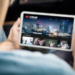 Personne tenant une tablette affichant un site Web de streaming vidéo automatique avec un arrière-plan de paysage urbain et des miniatures multimédias.