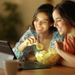 Deux personnes souriantes et mangeant des chips tout en regardant quelque chose sur une tablette à une table, avec l'auto Brouillon en arrière-plan ajoutant une touche supplémentaire d'excitation à leur décor décontracté.