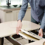 Une personne en chemise bleue rallonge une table extensible en y ajoutant une rallonge. La table est dressée avec des assiettes de nourriture et des parties de la cuisine de la maison sont visibles en arrière-plan.