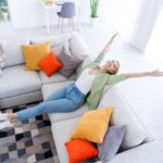 Une femme en tenue décontractée s'étire joyeusement sur un canapé en L aux coussins colorés dans un salon lumineux et moderne, ressemblant à l'esprit insouciant d'une auto à brouillon.