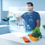 Un homme interagit avec un écran holographique dans une cuisine moderne. À côté du bol de légumes et d'une serviette sur le plan de travail, son automate Brouillon s'adapte parfaitement à chacune de ses commandes.