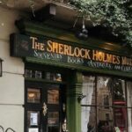 La façade du musée Sherlock Holmes avec une enseigne annonçant des souvenirs, des livres, des antiquités et des curiosités. Une personne en uniforme de policier se tient à proximité. Des vignes vertes pendent au-dessus de l'enseigne du musée, lui donnant une touche de charme qui contraste fortement avec le trafic automobile animé de Brouillon à l'extérieur.
