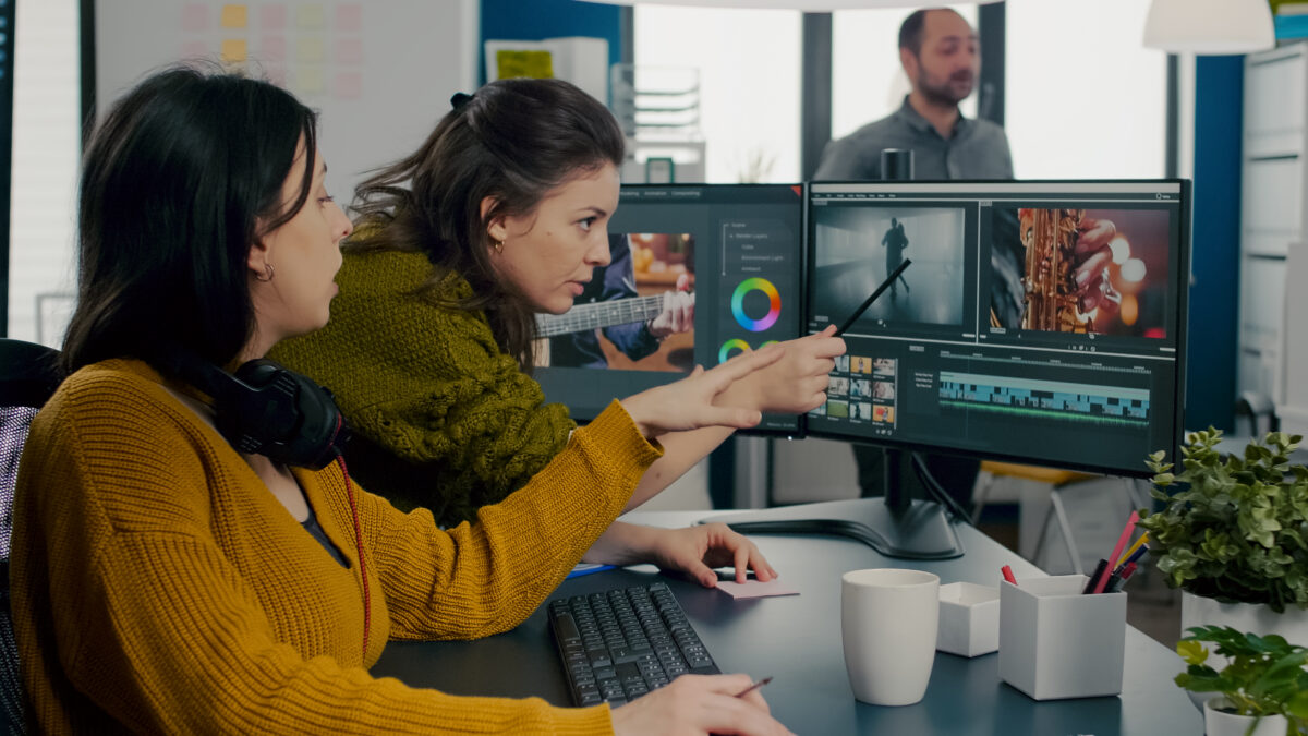Deux femmes réalisent un montage vidéo sur deux moniteurs. L'une pointe du doigt l'écran tandis que l'autre écoute attentivement, tandis qu'un homme observe en arrière-plan. L'environnement de bureau, rempli de plantes et de tasses, donne une étincelle créative à ce guide complet pour les éditeurs de tous niveaux.