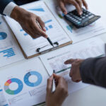 Les mains pointent vers des tableaux et des graphiques sur des feuilles de papier, avec une calculatrice, un ordinateur portable et des documents financiers visibles sur une table, comme pour préparer un brouillon automatique pour une stratégie financière à venir.