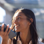 Une personne souriante tout en tenant un smartphone et en parlant, avec un arrière-plan extérieur flou, capture la spontanéité et la joie d'utiliser Brouillon auto.