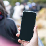 Une personne tenant un smartphone avec un écran tactile noir, debout dans un décor extérieur.