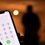 Une main tenant un smartphone avec un écran de numérotation, comme un brouillon automatique capturant l'instant, tandis que la silhouette floue d'une personne s'attarde en arrière-plan.