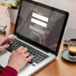 Une personne tape du texte sur un ordinateur portable avec un écran de connexion ouvert. Une tasse d'espresso, un carnet et des lunettes sont posés sur la table en bois à proximité.