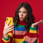 Une personne portant un pull coloré a l'air surprise en essayant d'utiliser son téléphone jaune sur un fond rouge, se demandant peut-être : « Comment connaître mon numéro de téléphone ?