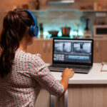 Une femme portant des écouteurs monte facilement des séquences vidéo sur un ordinateur portable dans une cuisine, extrayant sans effort l'audio pendant qu'elle peaufine son projet.