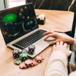 Une personne est plongée dans une partie de poker automatique Brouillon en ligne sur un ordinateur portable, entourée de plusieurs piles de jetons de poker colorés, assise à une table en bois poli.