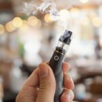 Une main tenant un stylo vape noir émettant de la fumée, avec un décor intérieur flou et un éclairage chaleureux en arrière-plan.