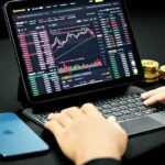 Une personne utilise un clavier avec une tablette affichant une plateforme de trading de crypto-monnaie et la fonction automatique Brouillon ; un smartphone et des pièces empilées sont sur la table.