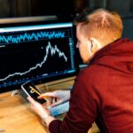 Une personne portant un sweat à capuche marron analyse le Forex et les crypto-monnaies, en regardant un graphique financier sur un écran d'ordinateur tout en utilisant un smartphone et une calculatrice à un bureau.