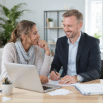 Un homme en costume et une femme en tenue décontractée sont assis à un bureau et discutent de documents relatifs au prêt immobilier pour 2025 tout en utilisant un ordinateur portable dans un bureau.