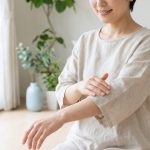 Une personne en vêtements clairs est assise à l'intérieur et se masse doucement l'avant-bras, peut-être après une séance d'épilation définitive, avec un salon lumineux et des plantes en arrière-plan.