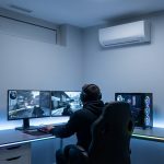 Personne assise à un bureau avec deux moniteurs, jouant sur un PC avec un éclairage RGB vibrant dans une pièce minimaliste et climatisée, tout en bricolant les derniers réglages automatiques de son Brouillon.